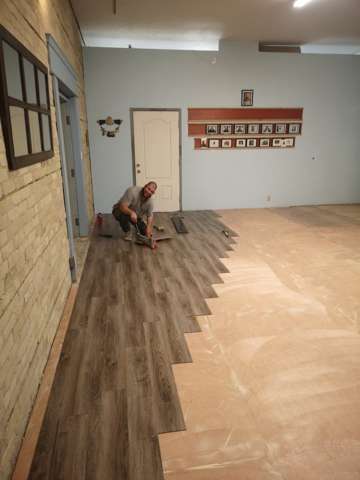 Photo shows Ark Aid male Volunteer Installs the New Beckham Brothers Reading Loose Caboose Luxury Vinyl Planks on sanded sub floor. Room has blue walls and Ark Aid memorabilia decorations on the walls.